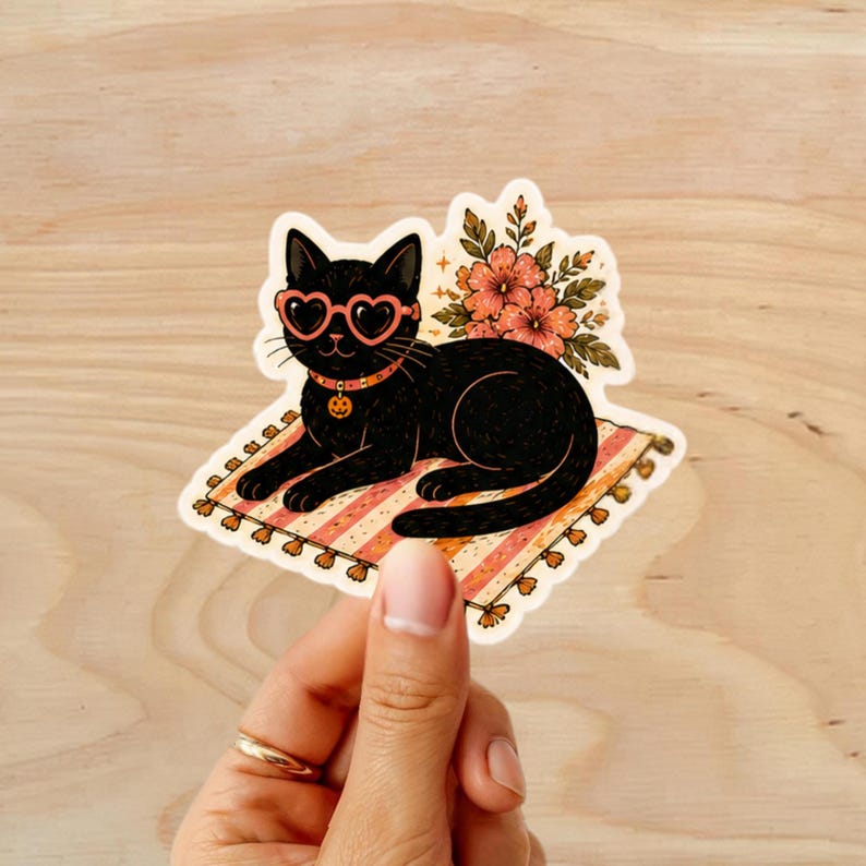 Puede incluir: Una pegatina con un gato negro con gafas de sol en forma de coraz&oacute;n y un collar de calabaza, tumbado en una alfombra a rayas. Hay flores detr&aacute;s del gato. La pegatina est&aacute; sobre una superficie de madera.