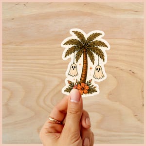 Puede incluir: Una pegatina con una palmera con dos fantasmas colgando. El &aacute;rbol tiene un tronco marr&oacute;n y hojas verdes, con flores naranjas y hojas verdes en la base. La pegatina est&aacute; sujeta por una mano.