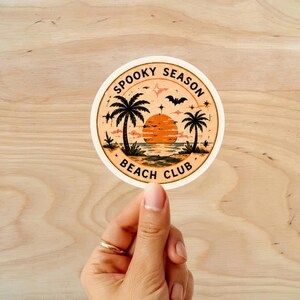 Puede incluir: Una pegatina redonda con una escena de playa, con palmeras, una puesta de sol y un murci&eacute;lago. El texto "Spooky Season Beach Club" est&aacute; impreso alrededor del borde. La pegatina est&aacute; sujeta por una mano.