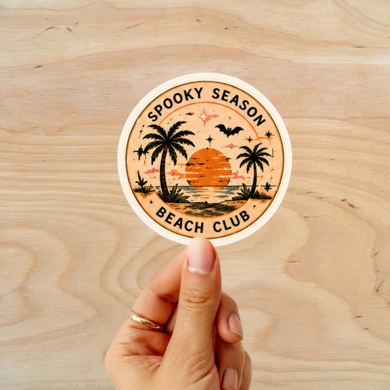 Puede incluir: Una pegatina redonda con una escena de playa, con palmeras, una puesta de sol y un murci&eacute;lago. El texto "Spooky Season Beach Club" est&aacute; impreso alrededor del borde. La pegatina est&aacute; sujeta por una mano.