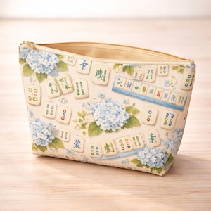 Op de afbeelding: Crèmekleurige make-uptas met ritssluiting en een bloemen- en Mahjong-tegelpatroon. Het ontwerp toont blauwe hortensia's en diverse Mahjong-tegels. De tas heeft een goudkleurige ritssluiting en een lichtgekleurde binnenkant.