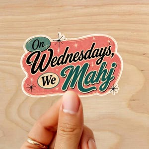 Może przedstawiać: Naklejka w stylu retro z napisem "On Wednesdays, We Mahj" w czarnych i turkusowych literach. Naklejka ma r&oacute;żowe i białe tło z akcentami w kształcie gwiazd. Naklejka jest trzymana przez osobę.