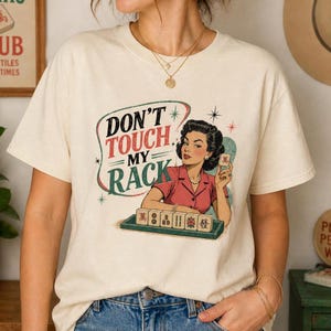 May include: Cream-colored t-shirt with a retro graphic. The design features a woman in a red top and the text "DON'T TOUCH MY RACK". The graphic includes starbursts and a tray of tiles.