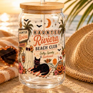 May include: A clear glass tumbler with a bamboo lid and straw, featuring a "Haunted Riviera Beach Club" design. The design includes a black cat wearing heart-shaped glasses, bats, palm trees, and a crescent moon. The tumbler is decorated with spooky beach-themed graphics.