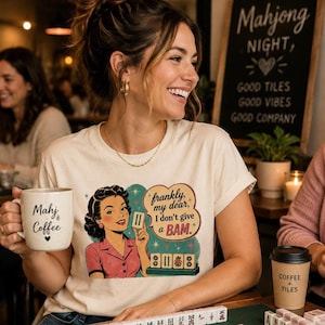 May include: A cream-coloured t-shirt with a retro illustration of a woman holding a Mahjong tile, with the text "frankly, my dear, I don't give a BAM." A coffee mug with "Mahj & Coffee" is held in the hand.