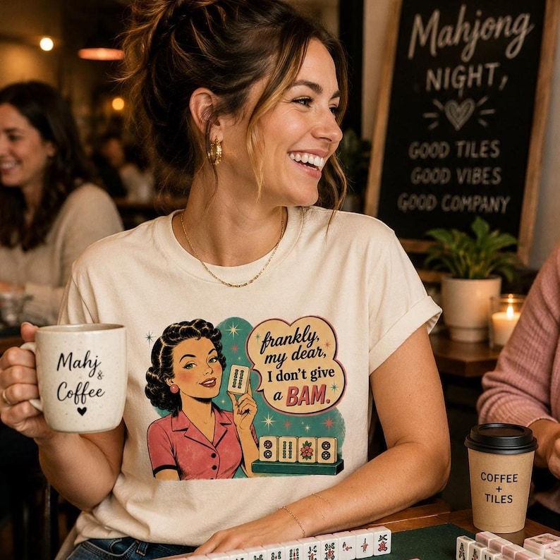 May include: A cream-coloured t-shirt with a retro illustration of a woman holding a Mahjong tile, with the text "frankly, my dear, I don't give a BAM." A coffee mug with "Mahj & Coffee" is held in the hand.