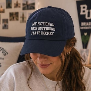Może przedstawiać: Granatowa czapka z daszkiem z białym haftowanym napisem z przodu o treści: "MY FICTIONAL BOOK BOYFRIEND PLAYS HOCKEY". Czapka posiada usztywnianą koronę, zakrzywiony daszek oraz guzik umieszczony na samym środku górnej części materiału.