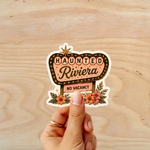 Puede incluir: Una pegatina con un letrero de estilo vintage que dice "HAUNTED Riviera" con una estrella y detalles florales. El letrero tiene un dise&ntilde;o retro con un borde negro y una pancarta "NO VACANCY".