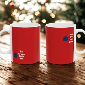 May include: Two white ceramic mugs with red exteriors and white handles. One mug has the text "be brave little dot" and a blue dot. The other mug has the text "you are not alone" and a blue dot. The mugs are on a wooden surface.
