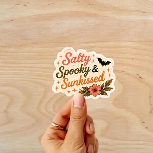 Puede incluir: Una pegatina con las palabras "Salty, Spooky & Sunkissed" en una fuente retro, con un murci&eacute;lago negro y un dise&ntilde;o floral. La pegatina est&aacute; sujeta por una mano con un anillo dorado, sobre un fondo de madera.