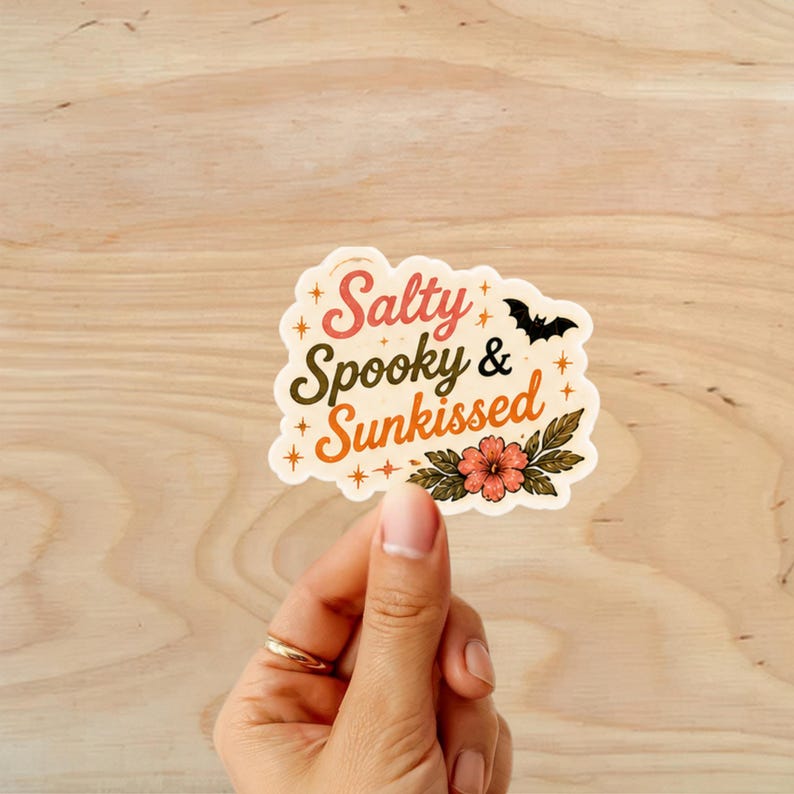 Puede incluir: Una pegatina con las palabras "Salty, Spooky & Sunkissed" en una fuente retro, con un murci&eacute;lago negro y un dise&ntilde;o floral. La pegatina est&aacute; sujeta por una mano con un anillo dorado, sobre un fondo de madera.