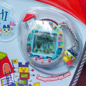 K&ouml;nnte beinhalten: Ein Tamagotchi-Spielzeug, ein virtuelles Haustier, in einer transparenten Kunststoffverpackung. Das Ger&auml;t hat einen blauen Bildschirm mit pixeligen Zeichen und einen gelben Anh&auml;nger. Der Text "Tamagotchieurope.com" ist sichtbar.