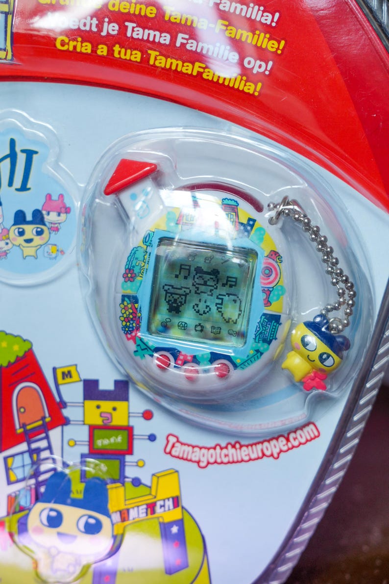 K&ouml;nnte beinhalten: Ein Tamagotchi-Spielzeug, ein virtuelles Haustier, in einer transparenten Kunststoffverpackung. Das Ger&auml;t hat einen blauen Bildschirm mit pixeligen Zeichen und einen gelben Anh&auml;nger. Der Text "Tamagotchieurope.com" ist sichtbar.