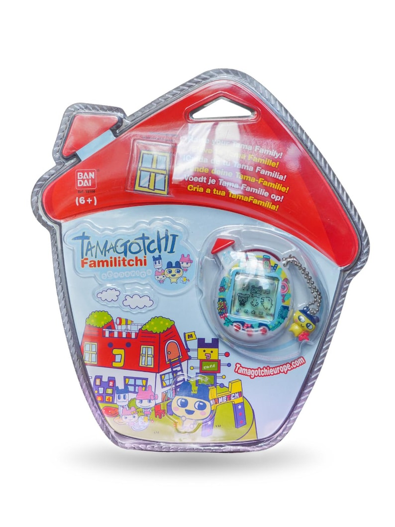 K&ouml;nnte beinhalten: Ein Tamagotchi Familitchi elektronisches Haustierspiel in einer hausf&ouml;rmigen Verpackung. Die Verpackung ist rot und aus transparentem Kunststoff, mit einer farbenfrohen Grafik der Tamagotchi-Charaktere und einer Stadtlandschaft. Das Spiel ist ein kleines Handheld-Ger&auml;t mit einem Bildschirm.
