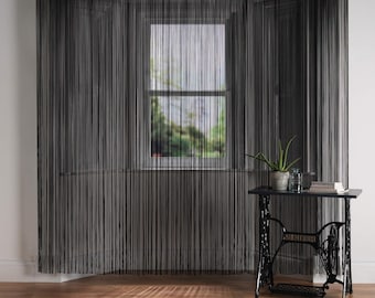 May include: Black string curtains hanging in a window. The curtains are made of thin strings that are tied together at the top. The window is behind the curtains and is out of focus. There is a small table with a plant on it in the foreground.