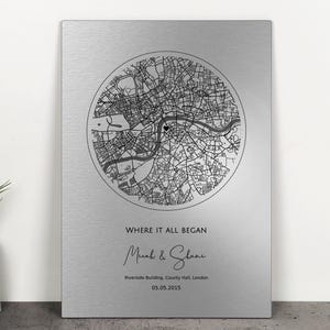 Puede incluir: Placa metálica plateada con un mapa en blanco y negro dentro de un círculo. El texto "WHERE IT ALL BEGAN" está debajo del mapa, seguido de nombres y una fecha. Un objeto decorativo, probablemente un regalo personalizado.