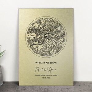 Puede incluir: Placa metálica dorada con un mapa en blanco y negro de una ciudad, rodeado. Debajo, el texto "WHERE IT ALL BEGAN" seguido de nombres y una fecha. La placa es rectangular y está en posición vertical.