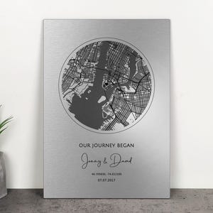 Puede incluir: Una pieza de arte mural de metal plateado con un mapa circular de una ciudad. El texto "OUR JOURNEY BEGAN" está sobre los nombres "Jenny & David" y una fecha y coordenadas. Una pieza decorativa para el hogar.