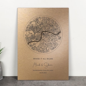 Pode incluir: Placa retangular dourada com um mapa circular detalhado de uma cidade. O texto "WHERE IT ALL BEGAN" está acima dos nomes "Micah & Shane" e da data "05.05.2015". O mapa é preto.