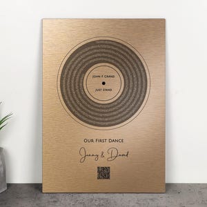 May include: A gold-colored plaque with a circular design resembling a vinyl record. The text "JOHN F GRAND" and "JUST STAND" are in the center. Below, it reads "OUR FIRST DANCE" with the names "Jenny & David" and a QR code.