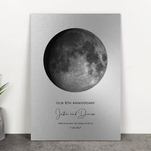 May include: A silver metal plaque featuring a detailed grayscale image of the moon. The text "OUR 8TH ANNIVERSARY" is above the names "Justia and Denise" in cursive. Additional text includes an address and date.