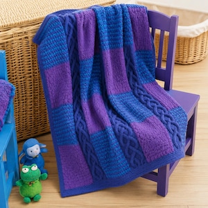 May include: A knitted blanket in shades of blue and purple, draped over a purple chair. The blanket features a cable knit design and color-blocked sections. A blue toy lamb and a green toy frog are on the floor.
