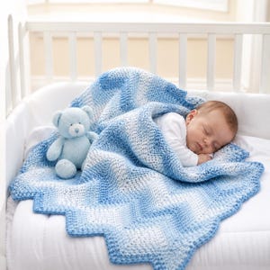 May include: A sleeping baby is nestled under a blue and white chevron-patterned crochet blanket. A light blue teddy bear sits beside the baby. The blanket has a scalloped edge and is draped over a white crib.