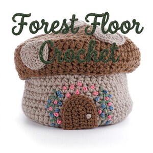 May include: A crocheted mushroom house in shades of brown, tan, and beige. The mushroom cap is brown with tan accents, and the base features a small brown door and floral embroidery. The text "Forest Floor Crochet" is above the mushroom.