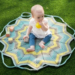 Peut inclure: Un bébé est assis sur une couverture crochetée en forme d'étoile. La couverture présente un motif en chevrons dans des tons de bleu, jaune et gris. Le bébé porte un haut blanc et un pantalon à pois bleus. Des blocs de bois sont éparpillés autour du bébé.
