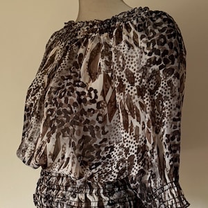 May include: A sheer, off-the-shoulder blouse with a brown and white animal print. The top features a gathered neckline, elasticated waist, and three-quarter sleeves with elastic cuffs. The fabric is lightweight and flowing.
