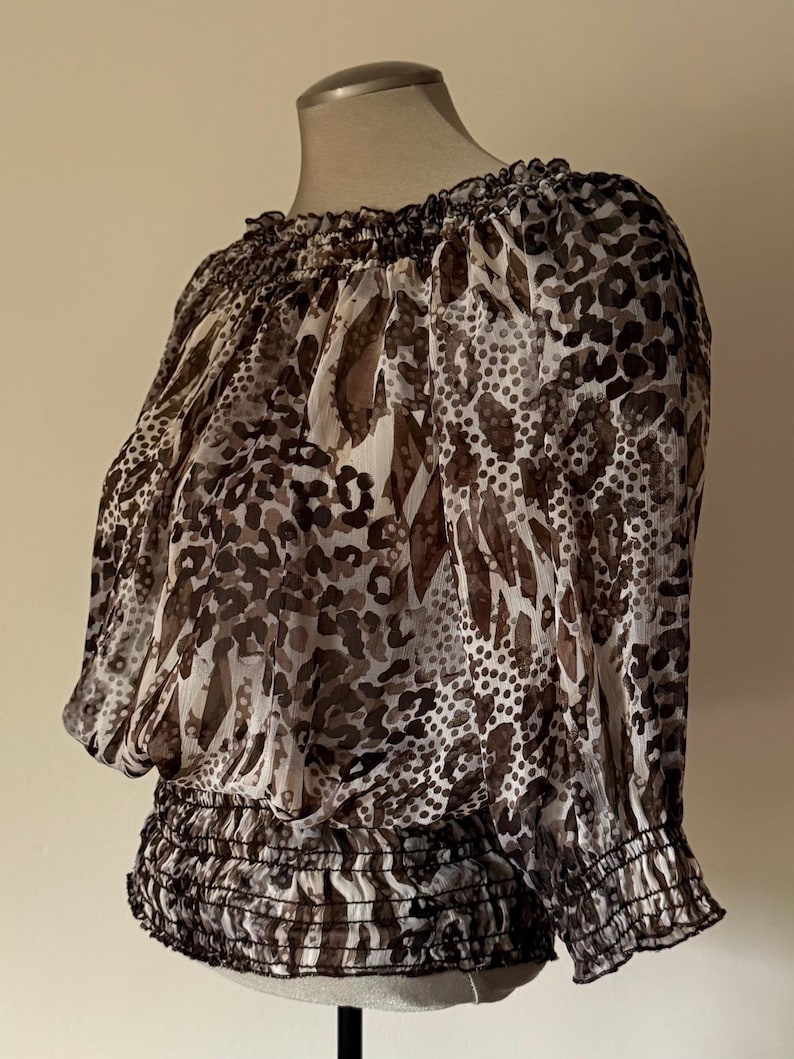 May include: A sheer, off-the-shoulder blouse with a brown and white animal print. The top features a gathered neckline, elasticated waist, and three-quarter sleeves with elastic cuffs. The fabric is lightweight and flowing.