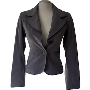 Könnte beinhalten: Ein grauer Blazer mit einem Ein-Knopf-Verschluss und fallenden Revers. Die taillierte Jacke hat lange Ärmel und ein klassisches Design, geeignet für professionelle oder halbformelle Anlässe. Der Blazer wird auf einer Schaufensterpuppe präsentiert.