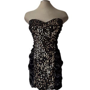 May include: A strapless black mini dress with a sweetheart neckline. The bodice is covered in silver sequins, and the sides feature gathered black satin. The dress is displayed on a mannequin.