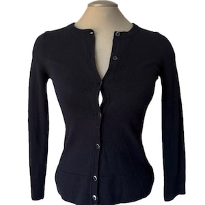 May include: A dark navy blue cardigan sweater with long sleeves and a button-up front. The sweater is displayed on a mannequin and has a classic, fitted silhouette. The buttons are small and match the color of the sweater.