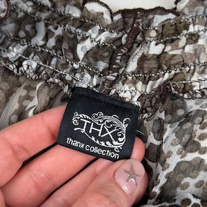May include: Close-up of a sheer top with a brown and white animal print pattern. The top has a ruffled neckline and a black tag that reads "THX thanx collection".