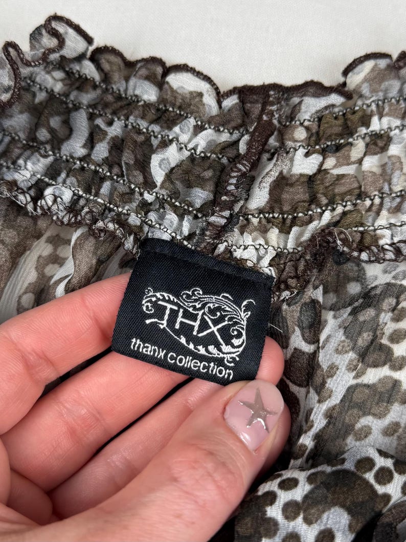 May include: Close-up of a sheer top with a brown and white animal print pattern. The top has a ruffled neckline and a black tag that reads "THX thanx collection".