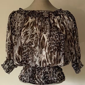 May include: A sheer, off-the-shoulder blouse with a brown and white animal print. The top features a ruffled neckline and three-quarter sleeves with elastic cuffs. The waistline is also elasticized.