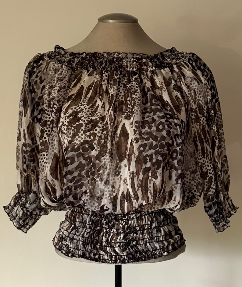 May include: A sheer, off-the-shoulder blouse with a brown and white animal print. The top features a ruffled neckline and three-quarter sleeves with elastic cuffs. The waistline is also elasticized.
