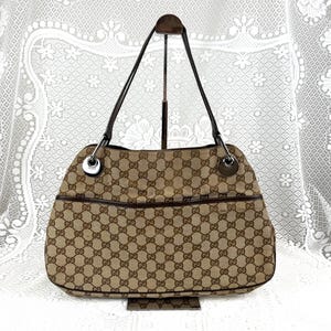 Puede incluir: Un bolso de mano de lona en color beige y marrón con el patrón de monograma GG de Gucci. El bolso cuenta con ribetes de cuero marrón oscuro, dos correas de hombro finas de cuero marrón oscuro y anillas de metal plateado en las asas.