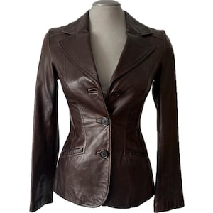 Puede incluir: Un blazer de cuero marrón oscuro con un corte entallado. La chaqueta presenta un cuello con solapas, dos bolsillos delanteros y cierre de tres botones. El blazer se exhibe en un maniquí sobre un fondo blanco.