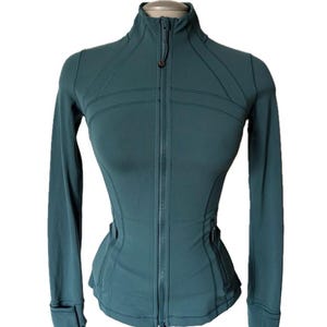 Può includere: Giacca sportiva color teal con cerniera intera e collo alto. La giacca a maniche lunghe ha una silhouette aderente con cuciture e dettagli di design sulle spalle e in vita. Sono presenti fori per i pollici sui polsini.