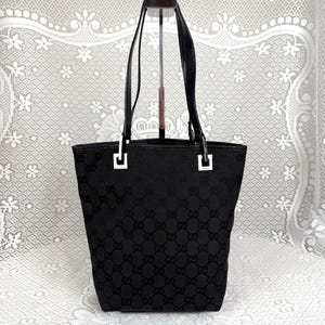 Puede incluir: Un bolso tote negro con un patrón de monograma GG tonal repetido en toda la superficie de tela. El bolso incluye dos correas negras para el hombro sujetas con herrajes metálicos de color plateado en las esquinas superiores y una base.