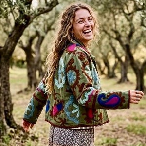 May include: A woman wearing a green jacket with colorful embroidered floral designs and a long, patterned skirt. The jacket has blue, pink, and brown accents. The skirt has a layered, bohemian style.