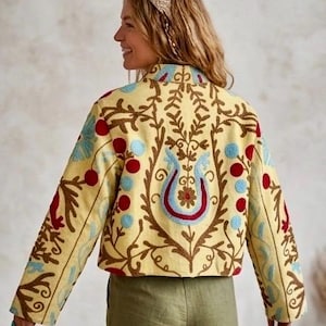 Pode incluir: Casaco amarelo bordado com um desenho floral em vermelho, azul e castanho. O casaco tem mangas compridas e gola redonda. O bordado apresenta um motivo floral central e padrões decorativos. O casaco é um estilo cropped.
