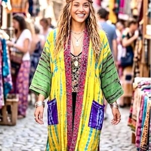 Könnte beinhalten: Eine Frau trägt einen langen Kimono im Batik-Stil in Gelb, Grün und Blau mit lila Taschen. Sie trägt ein schwarzes Oberteil und mehrreihige Halsketten. Der Hintergrund zeigt einen Markt mit bunten Textilien.