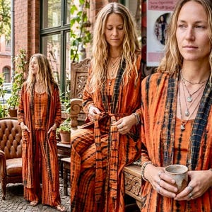 Può includere: Un completo tie-dye arancione e nero, composto da un lungo cardigan, un top e pantaloni. L'outfit è indossato da una persona con lunghi capelli biondi, accessoriato con collane e anelli. L'ensemble ha un'estetica bohémien.