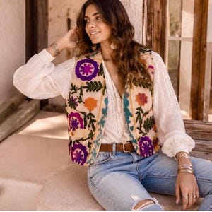 Puede incluir: Un chaleco sin mangas con base de color crema y bordados florales vibrantes en tonos morado, naranja y verde. El chaleco se lleva sobre una blusa blanca de manga larga con detalles de encaje, combinada con vaqueros azul claro y un cinturón marrón.
