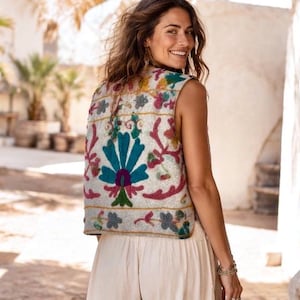 Puede incluir: Un chaleco sin mangas de color blanco roto con bordados florales y botánicos de colores vibrantes en tonos turquesa, magenta, verde y amarillo. El chaleco tiene un dobladillo recto y un diseño sencillo abierto por delante.