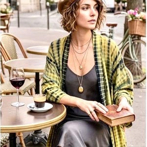 Puede incluir: Una mujer con boina marrón y botas largas marrones está sentada en una mesa de café, sosteniendo un libro. Lleva un vestido gris y un kimono con estampado amarillo y verde. Una copa de vino tinto y una taza de café están sobre la mesa.