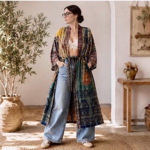 Könnte beinhalten: Ein langer, gemusterter Kimono in Gelb-, Grün- und Blautönen, getragen über einem weißen Oberteil und hellblauen Jeans. Der Kimono hat einen Bindegürtel und aufwendige Designs. Das Outfit wird mit Turnschuhen abgerundet.